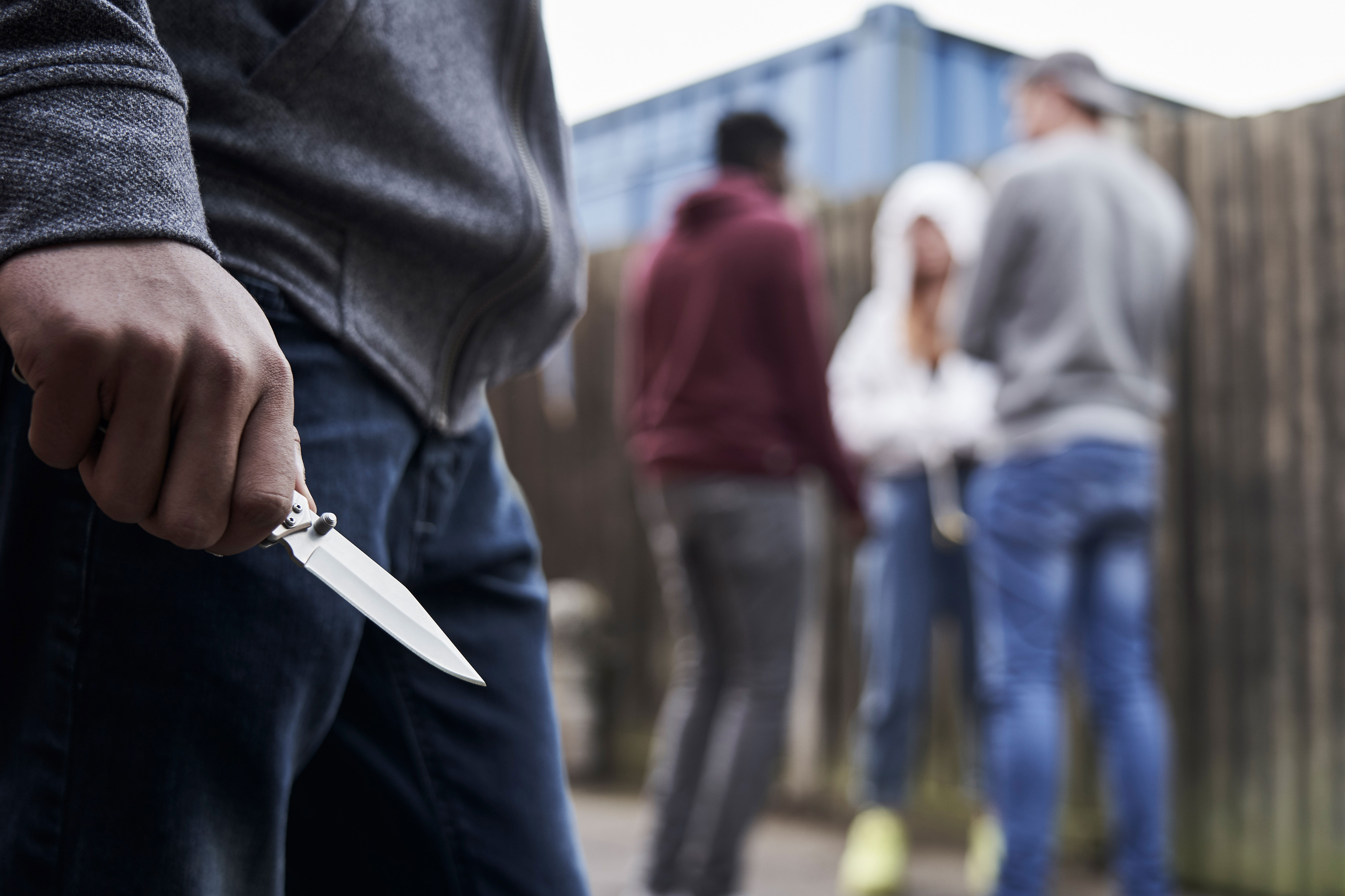 On the left, close-up of a knife in the hand of an unknown person, in the background a group of three people out of focus