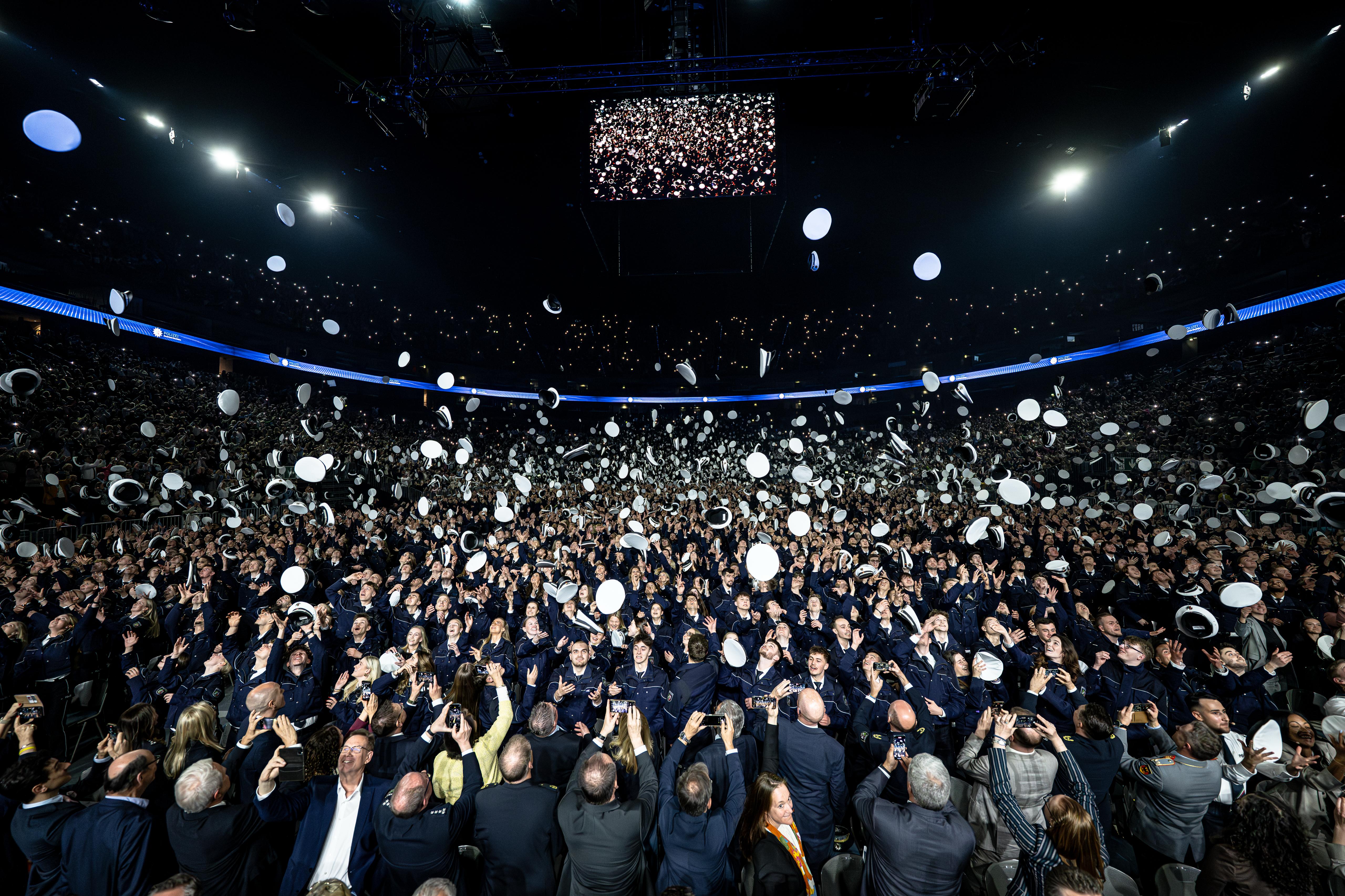Bei der Vereidigungsfeier in der Lanxess Arena in Köln werfen dreitausend Polizeianwärterinnen und -anwärter ihre Dienstmützen feierlich in die Luft. Sie tragen Uniform. Im Hintergrund ist es dunkel. Ein großer Bildschirm überträgt die Szene für das Publikum.