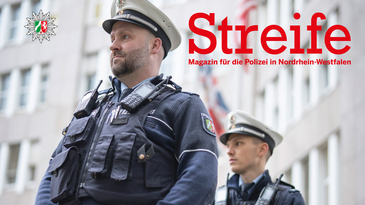Zwei Polizisten stehen vor dem britischen und dem US-amerikanische Generalkonsulat in Düsseldorf. Sie bewachen es. Beide tragen Uniform, ihre Blicke gehen zum linken oberen Rand des Bildes. Im Hintergrund ist verschwommen das Gebäude und Fahnen zu sehen.