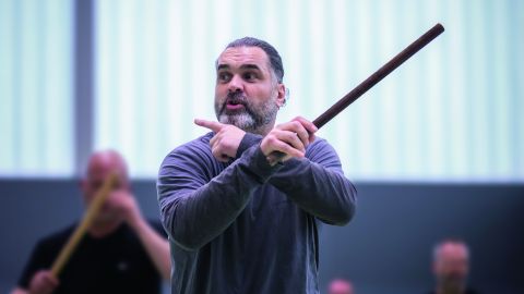 A trainer demonstrates movements with a baton in a training room. People can be seen blurred in the background.